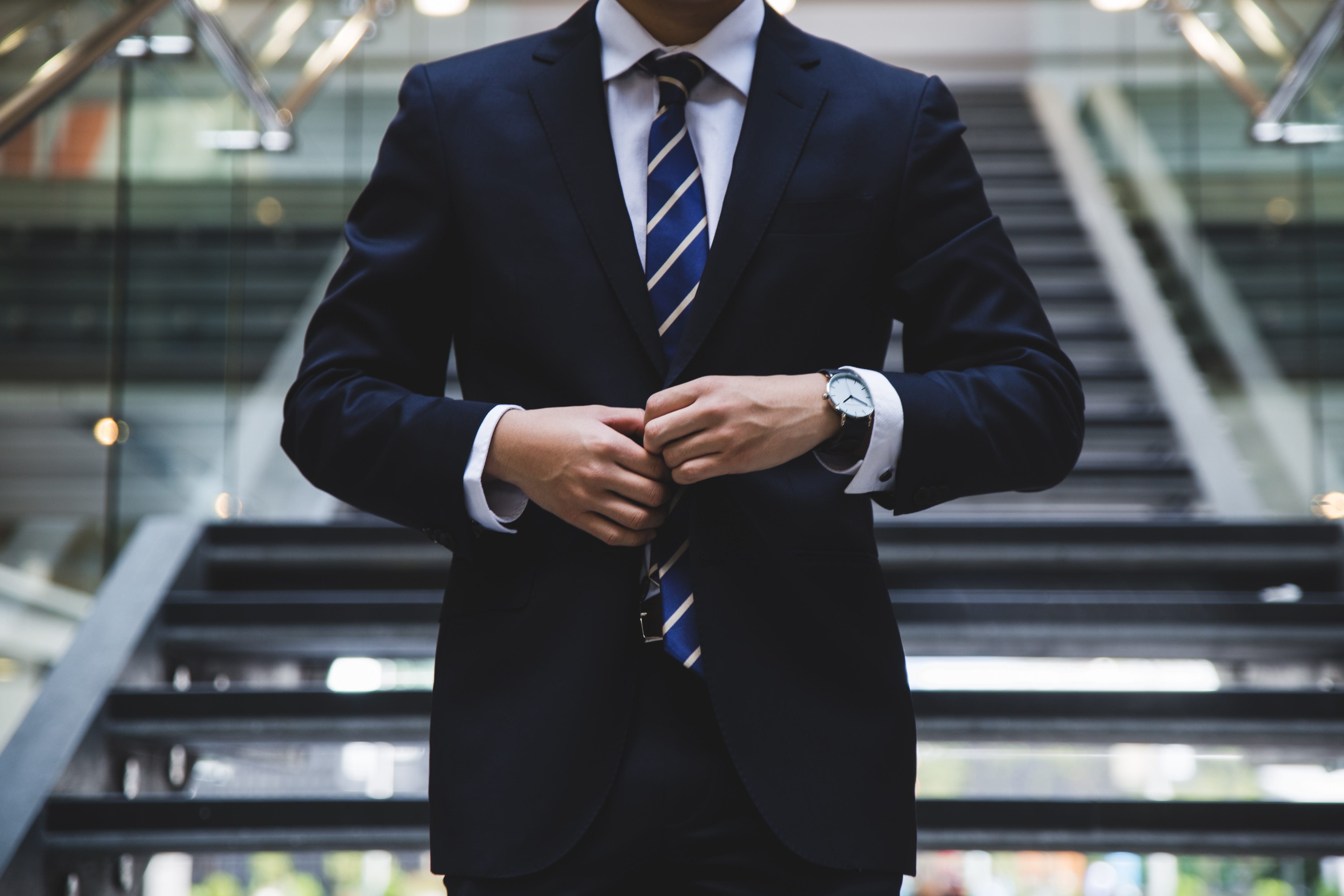Man in navy blue suit buttoning jacket while standing in front of stairs; image by Hunters Race, via Unsplash.com.