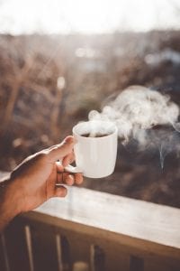 Man holding white ceramic cup with steaming hot coffee; image by Clay Banks, via Unsplash.com.
