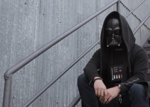 A dark-clad, hooded figure in a black mask sits on a metal stairway against a wall.