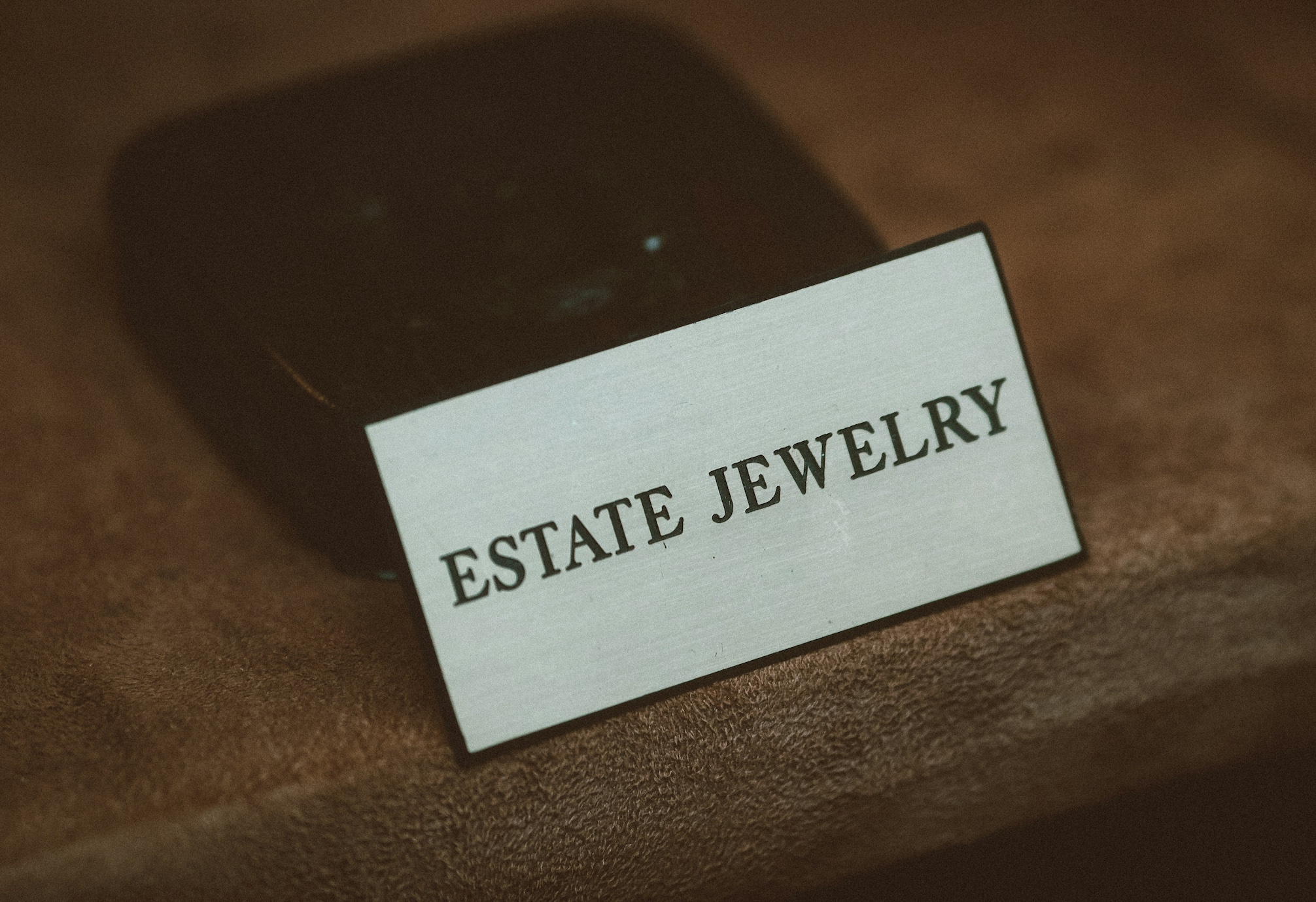 Metal sign saying Estate Jewelry leaning against jewelry box, all on brown background; image by Jon Tyson, via Unsplash.com.