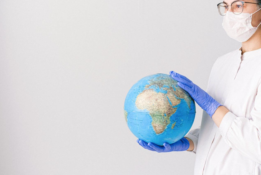 Person wearing face mask and lab coat holding a globe; image by Anna Shvets, via Pexels.com.