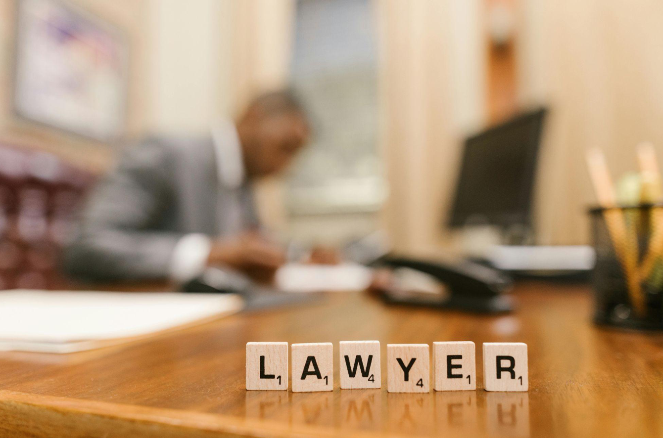 Close up of Scrabble letters spelling Lawyer on desk, man in background; image by RDNE Stock Project, via Pexels.com.