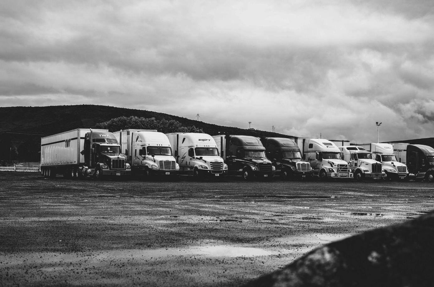 Greyscale photo of semi-trucks parked in a row under a cloudy sky; image by Kevin Bidwell, via Pexels.com.