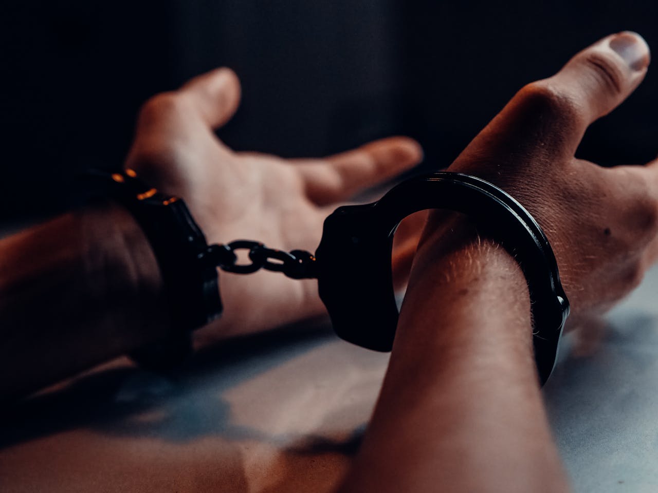 Person's hands in handcuffs with hands on table; image by Kindel Media, via Pexels.com.