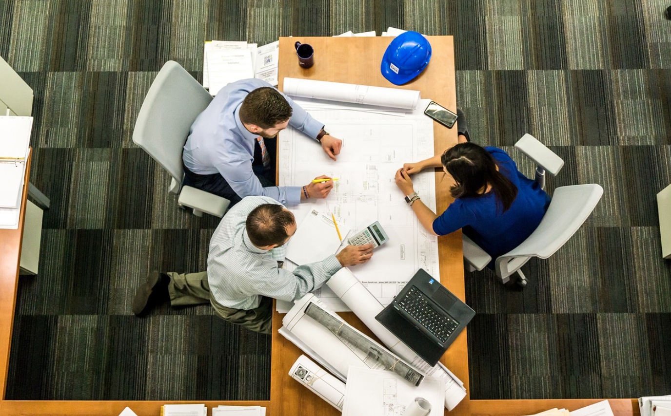 Overhead shot of two men and a woman review architectural drawings; image by mwitt1337, via Pixabay.com.