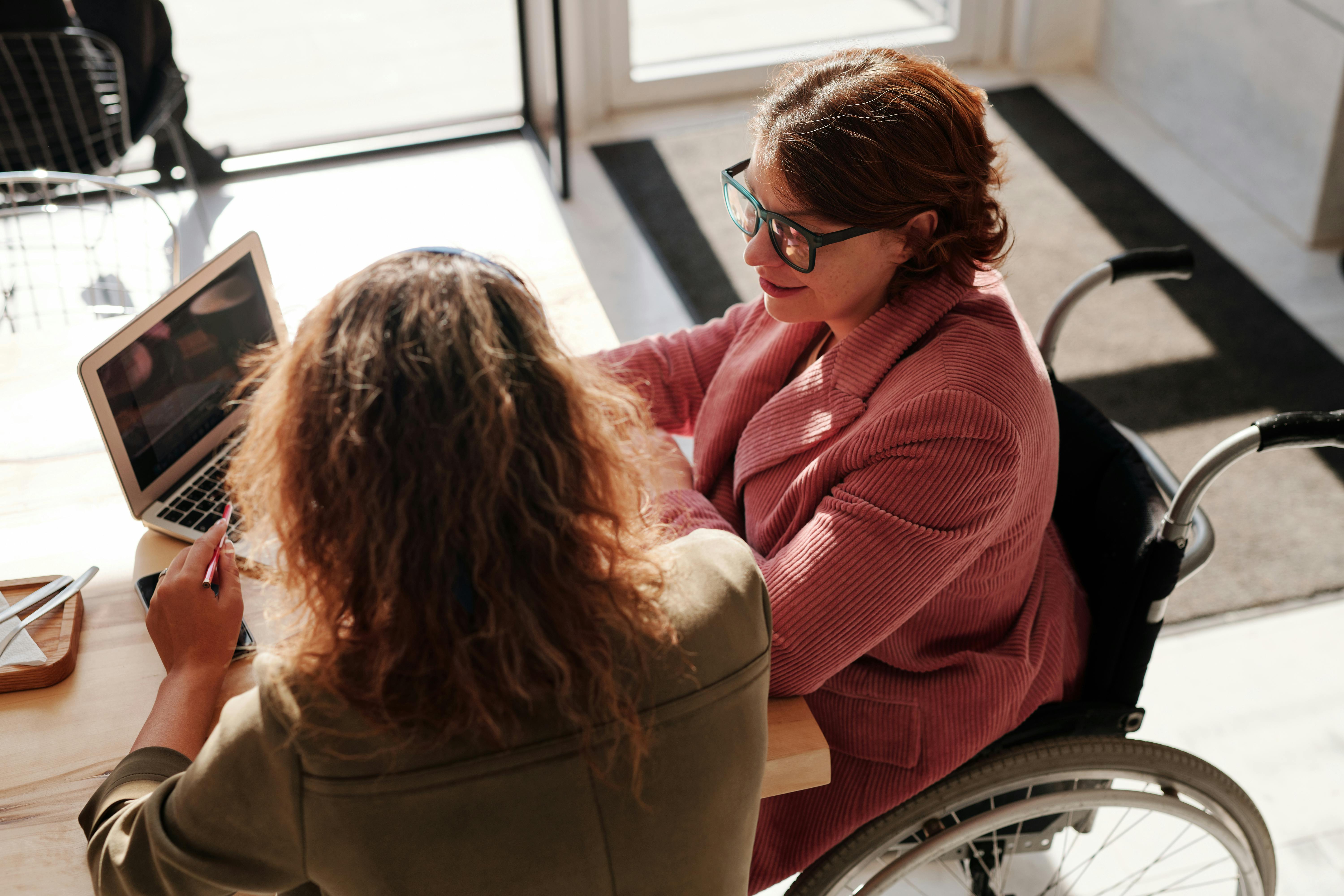 Woman in mauve blazer sitting in wheelchair at table with another woman; image by Marcus Aurelius, via Pexels.com.