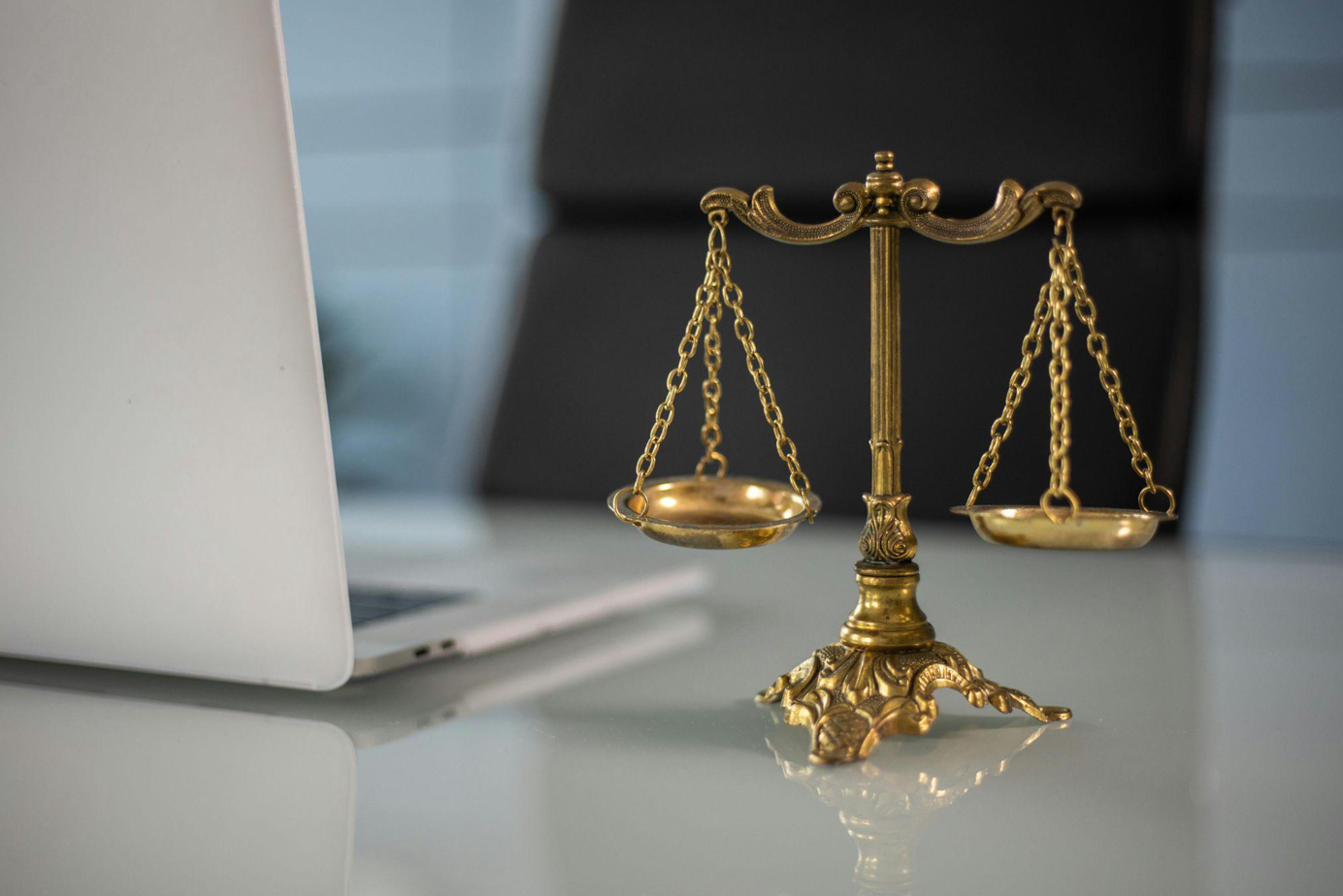 Scales of justice on desk by laptop; image by KATRIN BOLOVTSOVA, via Pexels.com.