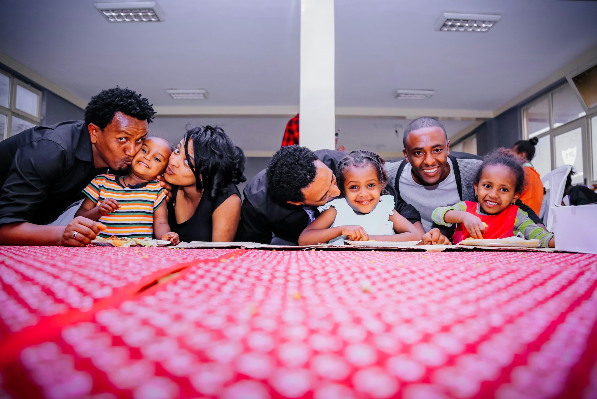 Woman and three men with three children on front of desk with red cloth; image by Gift Habeshaw, via Unsplash.com.