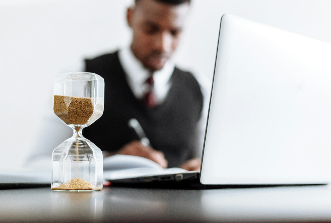 Selective focus shot of hourglass with man at desk with laptop in the background; image by Thirdman, via Pexels.com.