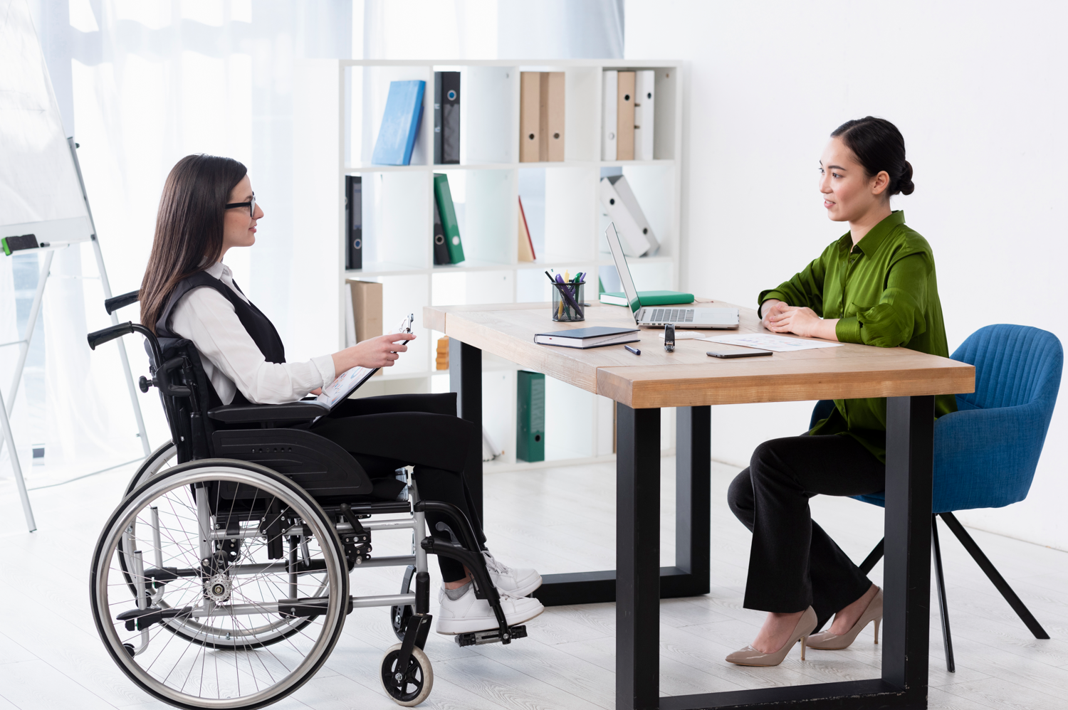 Woman at desk interviewing woman in wheelchair; image by Freepik, via Freepik.com.