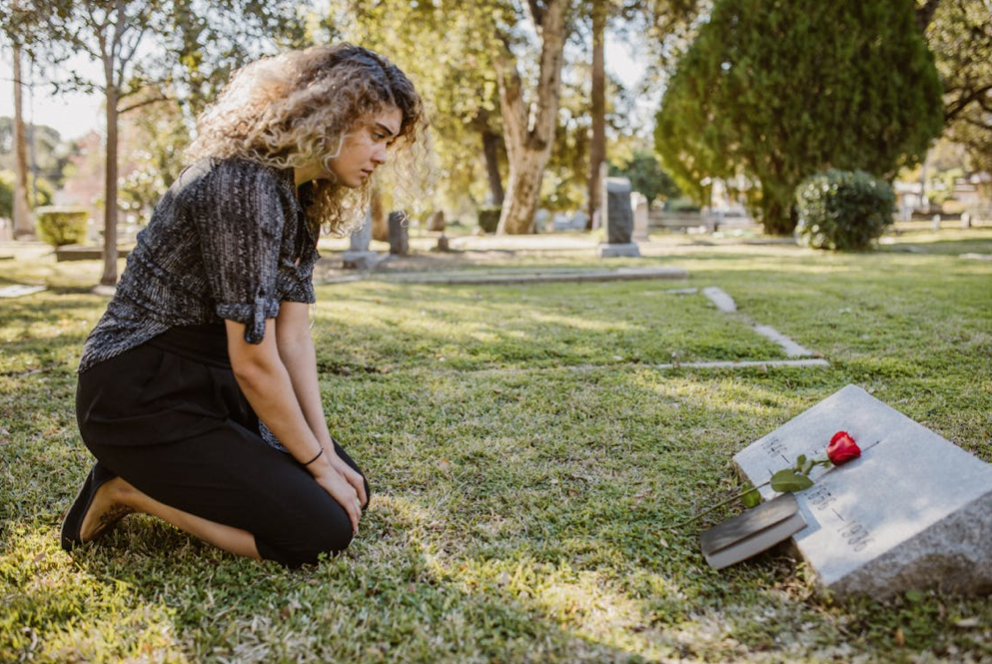 Woman kneeling at gravesite, a Bible and rose resting on headstone; image by RDNE Stock Project, via Pexels.com.